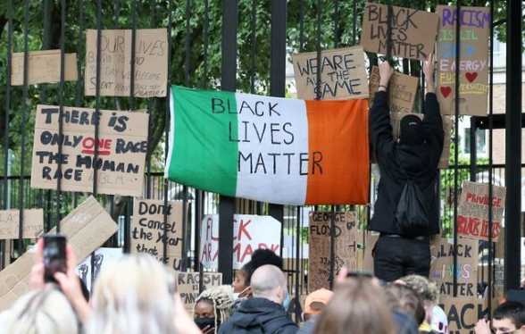 A protest scene featuring a person holding an Irish flag with the text 'BLACK LIVES MATTER' on it, surrounded by numerous signs advocating for racial justice.