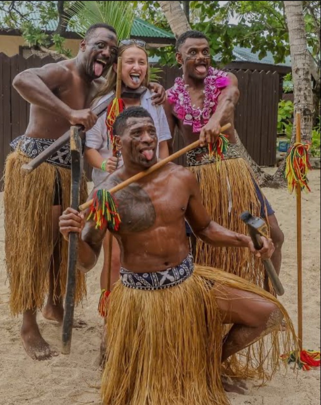 A group of four individuals, dressed in traditional attire featuring grass skirts, pose for a playful photo on a sandy beach. Two individuals are holding wooden sticks. They all have silly expressions, sticking out their tongues, with the female participant wearing sunglasses and a floral necklace.