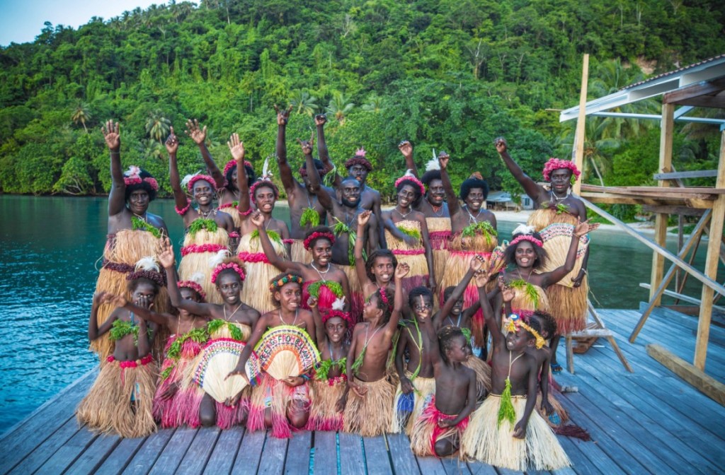 Group of Bougainvillean people wearing traditional attire, celebrating on a dock with a lush green background.