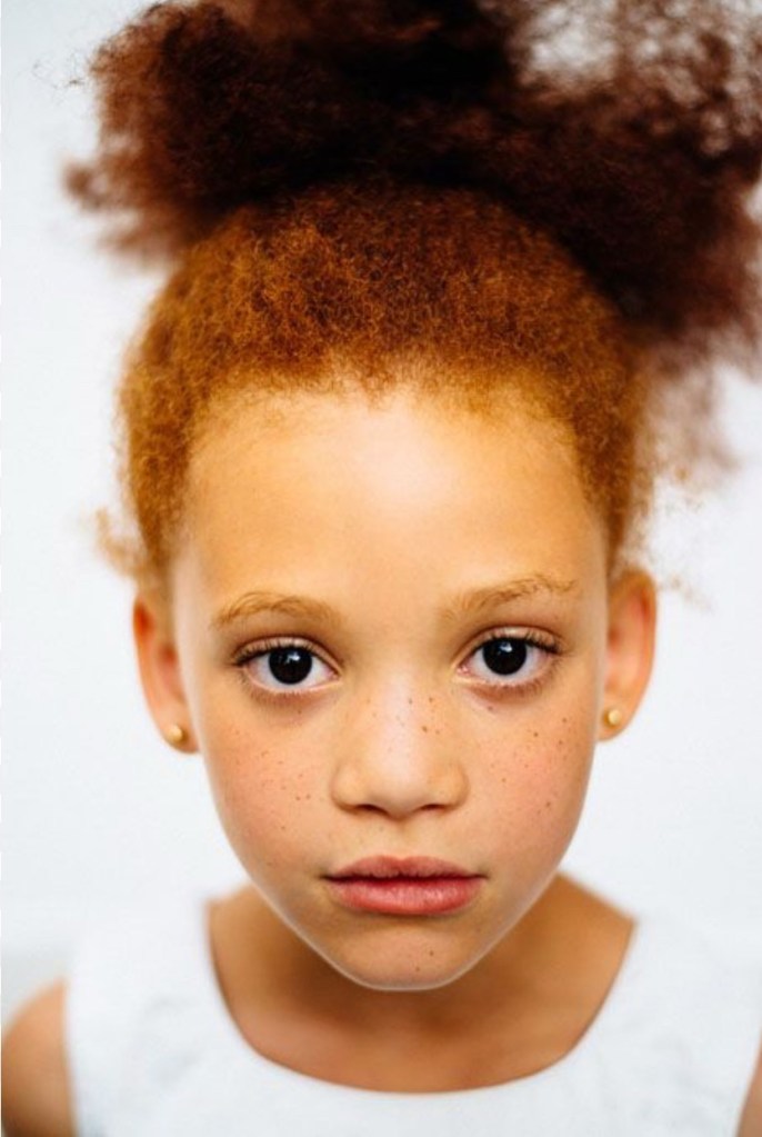 A close-up portrait of a young girl with red hair styled in a bun and freckles, looking directly at the camera.
