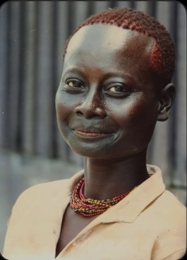 A smiling Black woman with short reddish-brown hair wearing a light-colored shirt and beaded necklace, against a blurred background.