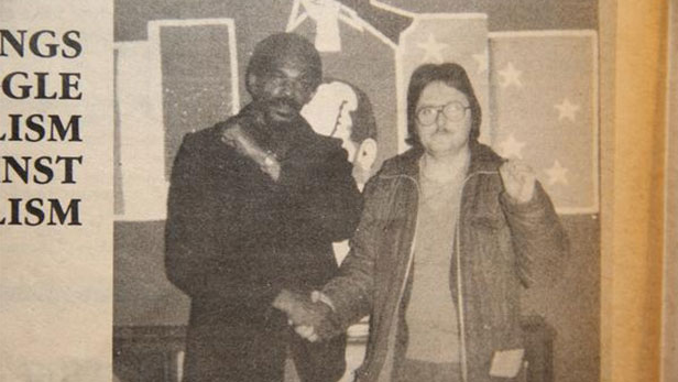 A man and a woman shake hands, symbolizing solidarity, in front of a backdrop featuring stars and political symbols.