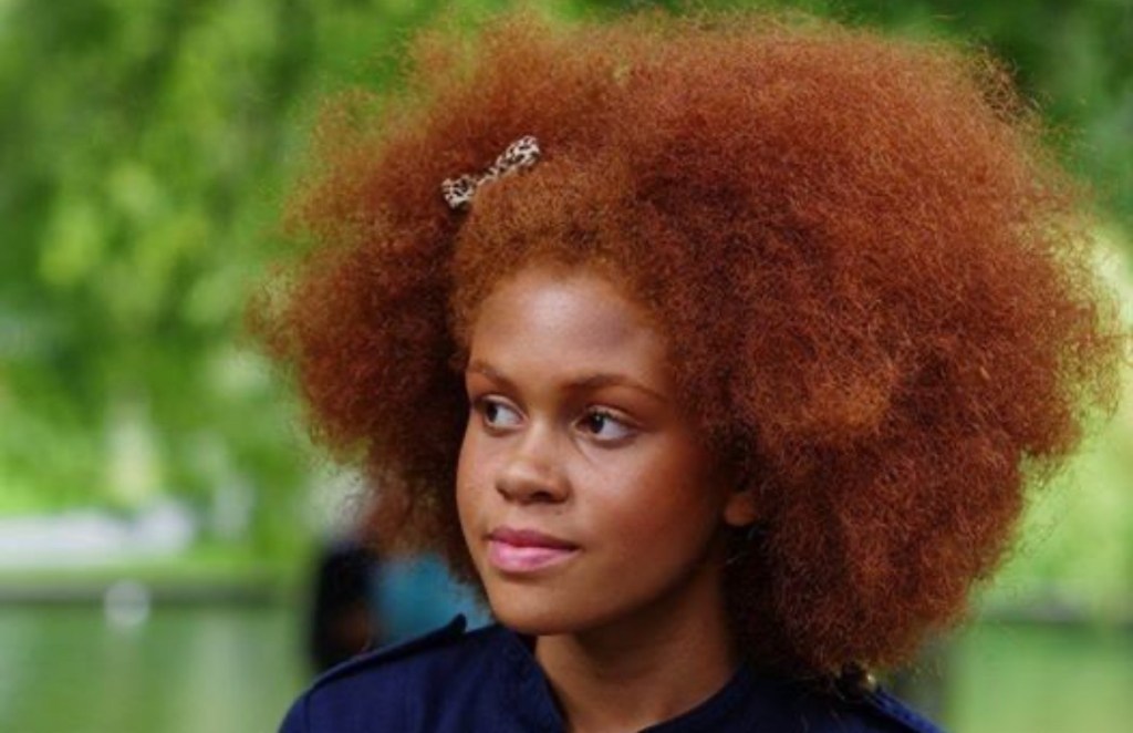 A young girl with vibrant red, curly hair, wearing a dark shirt, stands outdoors with a blurred green background.