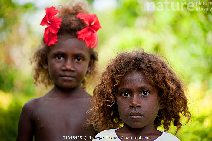 Two young girls with curly hair, one wearing red flowers in her hair, standing against a blurred green background.