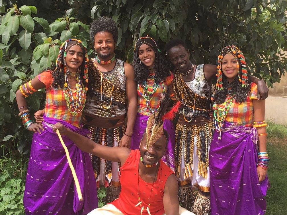 A group of six people dressed in colorful traditional attire, showcasing vibrant beadwork and cultural accessories, standing outdoors in front of green foliage.